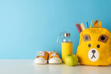 Okul temalı bir kompozisyonun yan görüntüsünde şirin bir sırt çantası, kırtasiye malzemeleri, spor ayakkabıları, su şişesi, mavi duvara dayalı elma. Eğitim tanıtımı ya da okula dönüş kampanyaları için mükemmel.
