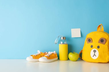 Sergilenen sınıf esasları: okul gereçleri, kalem tutacakları, renkli kalemler, cetveller, kitaplar, yapışkan notlar, plastikler ve elmalarla süslenmiş bir masanın yan görünüm fotoğrafı mavi bir arka fonda.