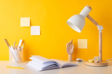 Akademik hazırlıklar. Kalemlerin, kalemlerin, cetvellerin, yapışkan bantların, açık hatırlatıcının, mankenin elinin, zımbanın, lambanın ve sarı duvara yapıştırılmış yapışkan notların yan görüntüsü.
