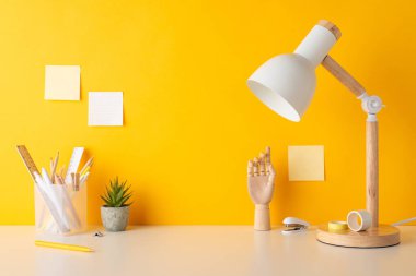 Akademik çalışma alanı kavramı. Kırtasiye malzemesi, kalem tutacağı, cetvel, kalem, yapışkan bant, manken el, zımba, saksı, lamba ve sarı duvardaki yapışkan notların yan görünümü