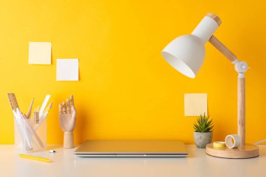 Çalışma ortamı konsepti. Kırtasiyeli masa resmi, tahta manken eli, saksı, dizüstü bilgisayar, lamba ve sarı duvara yapıştırılmış yapışkan notlar. Metin için kullanılabilir kopyalama alanı