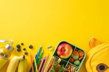 Dengeli okul yemeği konsepti. Sürdürülebilir beslenme çantasının üst görüntüsü taze, sağlıklı gıdalar, su şişesi, durağan, renkli kalemler, sarı arka planda canlı bir sırt çantası, mesaj veya reklam için yer.