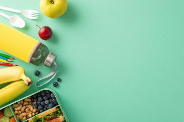 Okul gününü sağlıklı öğle yemeğiyle aydınlat: Doğal yiyecek, elma, erik, yabanmersini, kaju, sandviç, su şişesi, çatal bıçak, turkuaz arka planda renkli kalemler içeren plastik beslenme çantasının üst görüntüsü