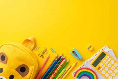 Eğitim kampanyaları için sarı izole edilmiş arka planda renkli okul kırtasiyeleriyle dolu sarı bir okul çantasının altını çizerek, etkili bir üst bakış açısıyla ebeveynleri ve eğitmenleri etkilemek