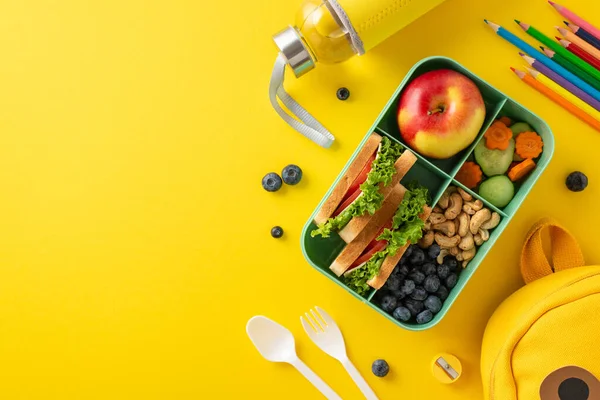 Nutritious school meal concept. Top view showing eco-conscious lunchbox containing organic treats, water bottle, cutlery, art supplies, playful backpack on yellow surface with room for text or promo