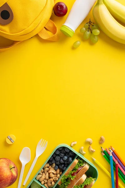 学校で生産的な日のために燃料 健康的な食べ物 ヨーグルト カトラリー カラー鉛筆 テキストや広告エリアで黄色の背景に面白いバックパックを誇るランチボックスの垂直トップビュー — ストック写真