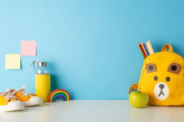 Okula dönüş titreşimleri: okul gereksinimlerinin, yapışkan notların, su şişesinin, elmanın, plastiğin, ayakkabıların, kırtasiyeli şirin sırt çantasının, mavi bir duvara yaslandığı bir yan görünüm fotoğrafı.