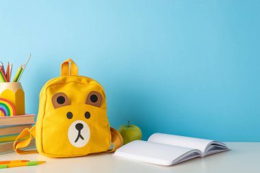 Malzemeler, kitaplar, elma, plasticin, kalem standı, çalışma kitabı ve mavi bir duvarda sevimli bir oyuncak ayı okul çantasıyla birlikte okul masasının yan görüntüsü. Okula dönüş promosyonları için ideal