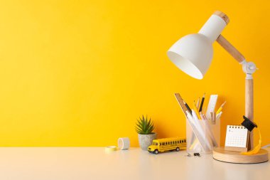 Akademik organizasyon kompozisyonu. Masa başı fotoğrafında kalemler, kırtasiye düzenleyicisi yöneticileri, takvim, akademik başlık, bitki, lamba, okul otobüsü oyuncakları, sarı duvar arkaplanında boş alan
