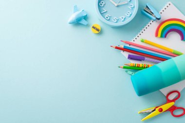 Yukarıdan bakıldığında iyi organize edilmiş çizim istasyonu: parlak kırtasiye, kalem kutusu, kalemler, eskiz defteri, plastik zımba, makas, uçak biçimli bileyici, yumuşak mavi zemin üzerindeki saat. Metin veya reklam için boşluk