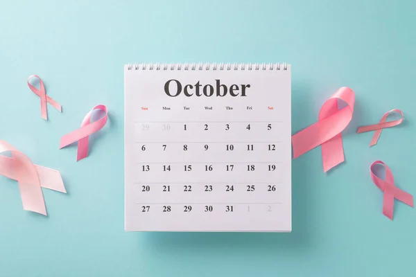 Breast cancer early detection arrangement. Top view shot of October calendar, surrounded by pink ribbons on pastel blue backdrop. Optimal for medical promotions