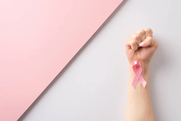 Commemorate International Breast Cancer Awareness Month with this top-view image of hand shaped into fist with pink ribbon on pink and white background. Perfect for text or advertising placements