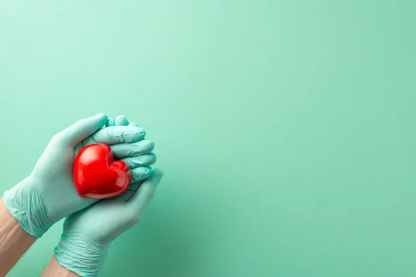 Safeguarding Against Heart Conditions. Aerial perspective picture displaying heart-embracing hands in medical gloves against a light teal isolated setting. Suitable area for text or adverts placement