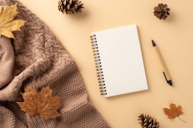 Sonbahar temalı ofis kurulumu: Sıcak örgü battaniye, not defteri, kalem, akçaağaç yaprakları ve çam kozalaklarının yukarıdan çekimi. Yumuşak bej arkaplan, mesajlaşma için mükemmel