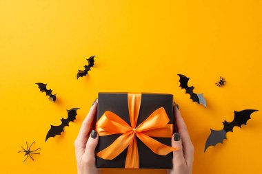 Cadılar Bayramını sevdiklerimiz için yükseltin. Perspektifin üstünde, siyah hediye kutusuyla bir kadının ellerini gösteriyor. Yay ve cadılar bayramı dekoru turuncu izole arka planda. Reklam ve mesajlara olanak sağlıyor.