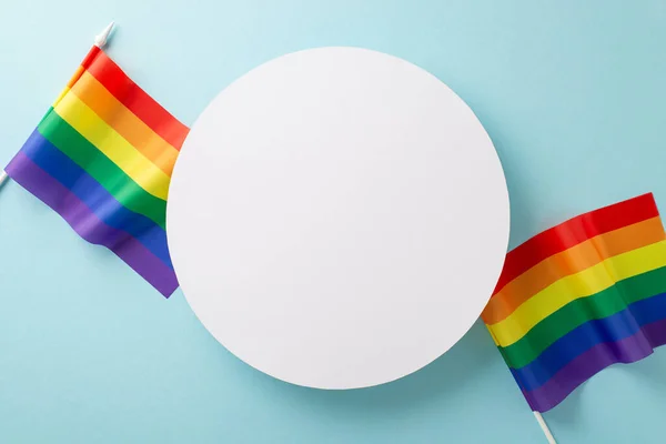 Commemorating LGBTQ+ Pride Month in July with pride flags around empty circle, displayed from above on a light blue isolated background, offering ample copy-space for ads or text integration