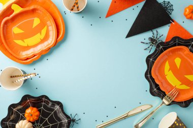 Büyüleyici bir Cadılar Bayramı partisi masası. Masa örtüsü, çatal bıçak takımı, kağıt bardaklar, kamışlar, çelenkler, konfeti ve diğer şenlik malzemelerini tanıtım için çerçeveyle birlikte pastel mavi arka planda sergiliyoruz.