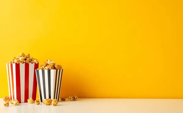 Movie night excitement! A side view of a table with cheese and caramel popcorn in striped boxes against a vibrant yellow wall with space for your message or promotion