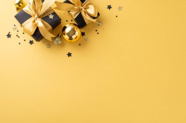 Noel ihtişamı motifi. Siyah hediye kutularının üst görüntüsü, zengin kurdele yayları, şık ağaç mücevherleri, ve sarı arka planda parlayan konfeti, mevsimlik metin veya promosyon için boş bir alan
