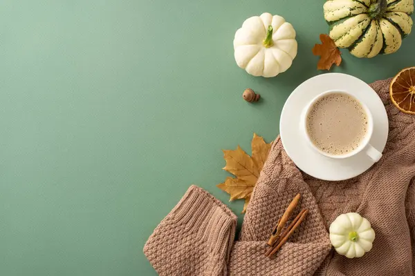 Embrace Autumn morning mood with top view shot showcasing warm knitted sweater, steaming cup of coffee, saucer, pumpkins, acorn, cinnamon sticks, other fall accents on green surface for text placement