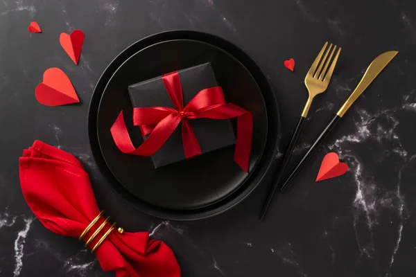 Dine-in restaurant on Valentine\'s Day theme. Top-view shot of beautiful giftbox in plate, hearts, elegant tableware, red napkin with ring on romantic black marble table setting, perfect for ad