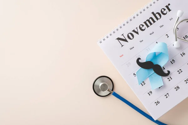 Raising awareness for Men\'s Health Month. Overhead image of blue ribbon, mustache silhouette, stethoscope, and November calendar on a pastel beige background, representing health checkup planning