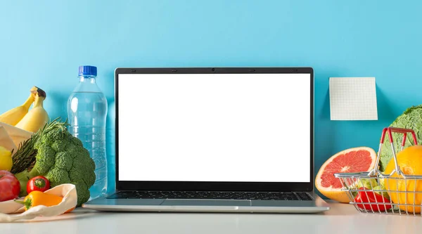 Fresh start to healthier you: On table, water bottle, veggies, fruits in market basket and bag, laptop for online shopping, sticky note with blue wall behind, perfect for adding text or advertisement
