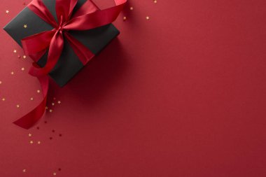 Lüks hediye: Kırmızı kurdeleyle süslenmiş, Marsala arka planında altın yıldız şeklindeki konfetilerin arasına yerleştirilmiş sofistike bir siyah hediye kutusunun üst görüntüsü.