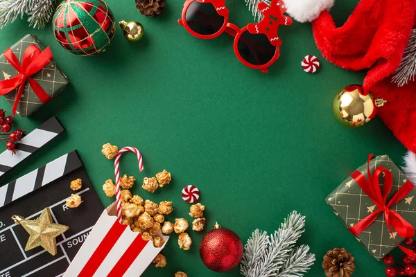 Welcome the New Year with movie night at cinema. Top view shot of clapperboard, funny glasses, popcorn in striped box, Santa\'s hat, baubles, star decor, candies, and frosty fir twigs on green backdrop