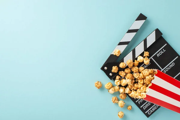 Filmstrip\'s grand premiere on horizon. Overhead image of movie producer\'s clapper and delectable popcorn scattered from striped container on light blue background, offering space for text or promotion