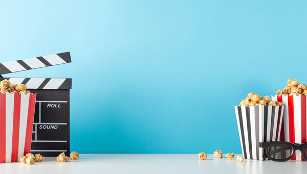 Elevate your film premiere using delightful snack motif. Capture side view of table featuring movie director's clapper, 3D specs, tasty popcorn against pastel blue backdrop, leaving space for movie ad