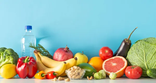 Nutrient-rich diet for a vibrant life concept. Side view shot of a table with water, veggies and fruits, such as cabbage, bell pepper, bananas, oranges, apple, and more, against a blue wall backdrop