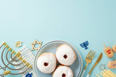 Hanukkah feast concept: top view of dreidel, doughnuts, fork, knife, napkin, candles, and Menorah on a pastel blue table setting. Perfect for ads clipart