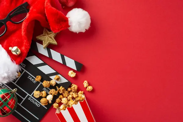 Celebrate the New Year at the movies. Top-down view of a clapperboard, 3D glasses, delicious popcorn in box, Santa\'s hat, baubles, sparkle star decor on red backdrop, empty space for text or promo
