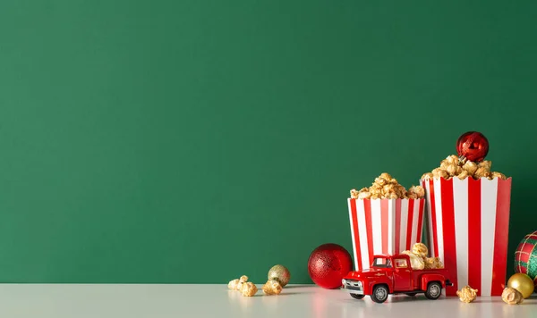 Create festive atmosphere for Noel with home popcorn delivery idea. Side view of tabletop, featuring charming red car, delectable popcorn, decorative balls. Green wall backdrop for movie ads or promo
