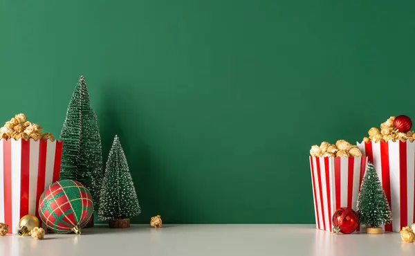 Christmas holiday kickoff promo idea. Side view shot of tabletop adorned with caramel popcorn, baubles and a miniature Christmas tree against a green backdrop for festive movie promotions
