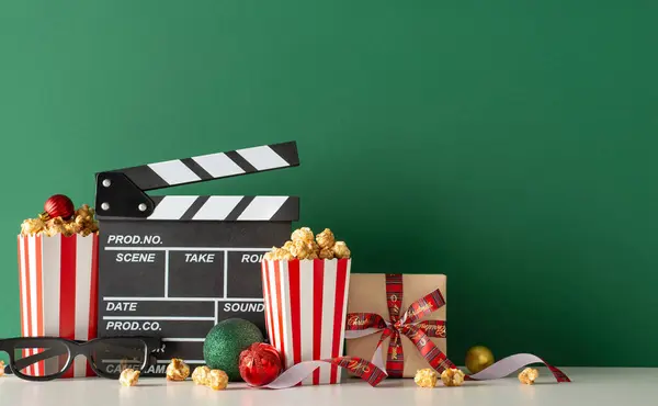Glamorous movie premiere with mouthwatering snacks motif. Side view table with movie slate, 3D glasses, popcorn, accompanied by festive balls and gift against green wall, offering canvas for movie ad