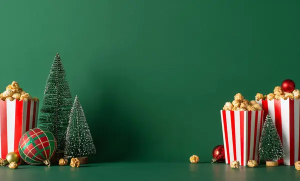 Yuletide movie premiere promo concept. Side view photograph of a table embellished with popcorn in striped buckets and petite evergreen tree against cozy winter-themed green background for ad space