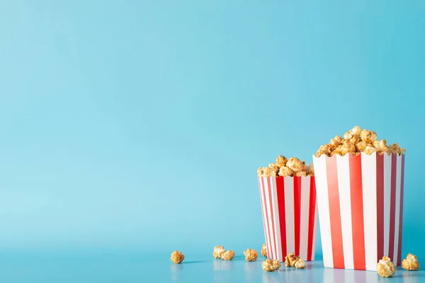 Cinema time with friends and snacks inspiration. Side view perspective picture of a tabletop filled with scrumptious popcorn in striped containers against a soft blue wall, great for movie promo