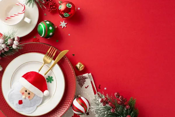 Craft delightful New Year\'s dinner table setup. Top view plates, charming gold cutlery in playful pocket, napkin, mug, candy cane, ornaments, frosted fir twigs, mistletoe on red backdrop with ad space