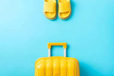 Seyahat zihniyetine ara ver. Sarı bir bavul ve parlak mavi bir yüzey üzerinde lastik parmak arası terlikler. Seyahat anlaşmalarınızı etkili bir şekilde tanıtın