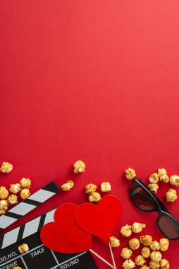 Sevgililer Günü 'nü romantik filmlerle ayarlayın: Bir klaket panosunun dikey üst görüntüsü, üç boyutlu gözlük, dağınık patlamış mısır ve kalp temalı dekor kırmızı arka planda, samimi bir film gecesi için ideal.