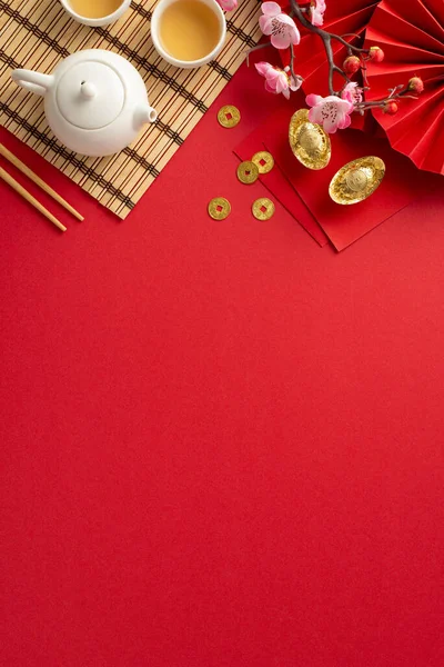 Opulent Chinese New Year table: Overhead vertical shot capturing traditional fan, tea set, chopsticks, bamboo mat, lucky coins, envelopes and sakura against red backdrop, leaving space for text or ads
