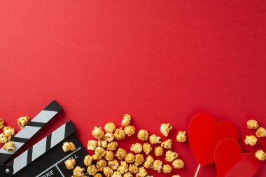 Romantik Film Gecesi: Kırmızı zemin üzerinde bir alkış tahtası, patlamış mısır ve kalp şeklindeki dekorun yukarıdan çekimi, aşk dolu bir film galası sahnesini hazırlıyor
