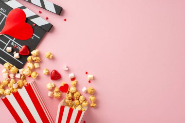 Sevgililer Günü 'nde romantizm var. Clapperboard, çizgili patlamış mısır kapları, kalp şeklindeki süsler, şekerler, şekerlemeler, tatlı pastel pembe yüzey üzerine reklam alanlarıyla serpiştirilmiş şekerlemeler