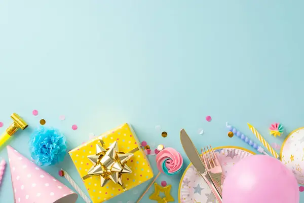 Colorful party setting idea. Overhead shot capturing festive table arrangement with paper plate, cutlery, sweets, gift, birthday headgear against pastel blue background, providing space for advert