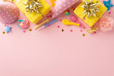 Charming birthday vignette concept. Top view of table decorated with adorable merengue lollipops, gift packages, birthday hats, and other joyful elements on pastel pink backdrop. Open area for text