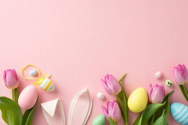 Paskalya temalı kompozisyonda canlı yumurtalar, sevimli tavşan kulakları ve laleler sergileniyor. Pastel pembe arkaplandaki üst görünüm, metin veya tanıtım içeriğiniz için hazır