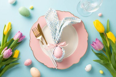 Tavşan temalı Zevk: Tavşan kulaklı peçete, tabak, çatal bıçak, şarap kadehi, laleler ve renkli yumurtalarla büyüleyici bir Paskalya masası manzarası. Pastel mavi arkaplan