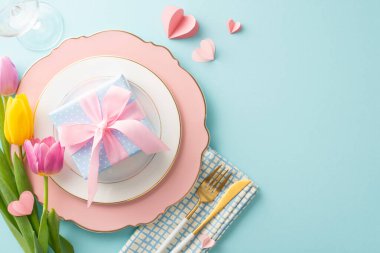 Zarif Kadınlar Günü masa düzenlemesi. Lüks tabağın, peçetenin, sofra takımının, şarap bardağının, nefis lalelerin, hediye kutusunun ve pastel mavi ayar üzerindeki kalplerin resmi. Mesaj ve reklamlar için boş bir alan.